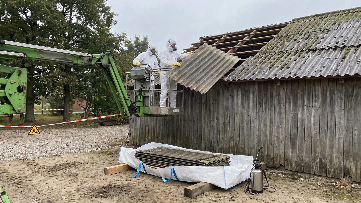 Zwei Arbeiter in weißen Schutzanzügen heben von der Hubarbeitsbühne aus eine Wellplatte aus Faserzement vom Dach, unten liegen ausgebaute Platten im Plattensack